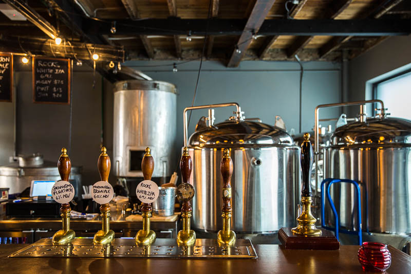 The interior of a craft brewery.  Several bar taps are positioned in front of two large fermentation tanks.